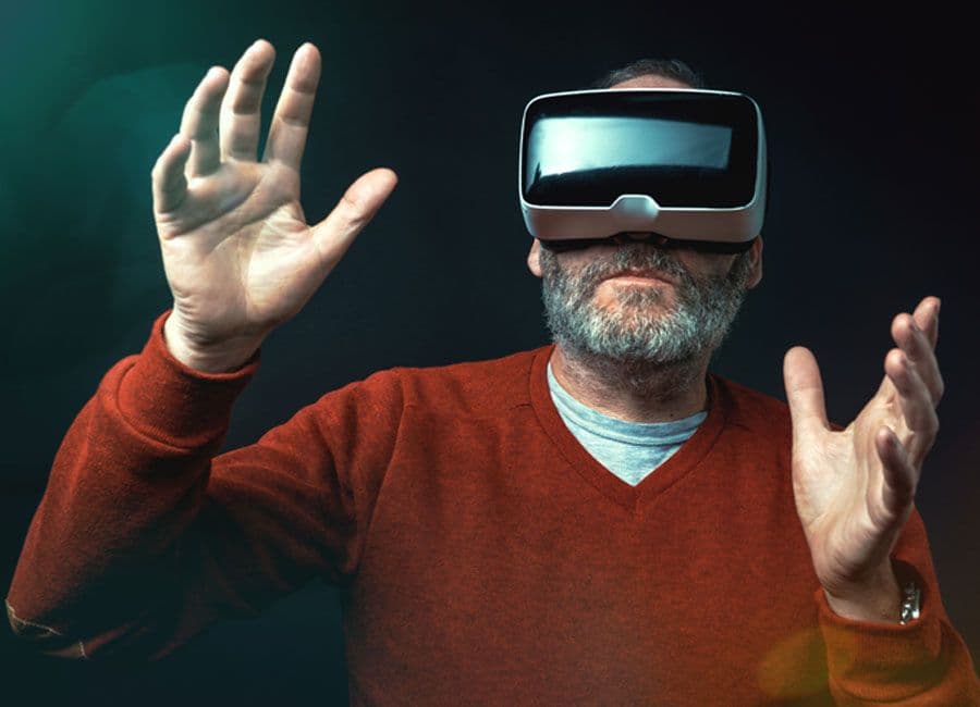 A man with a grey beard dons virtual reality glasses and holds his hands out in front of him.