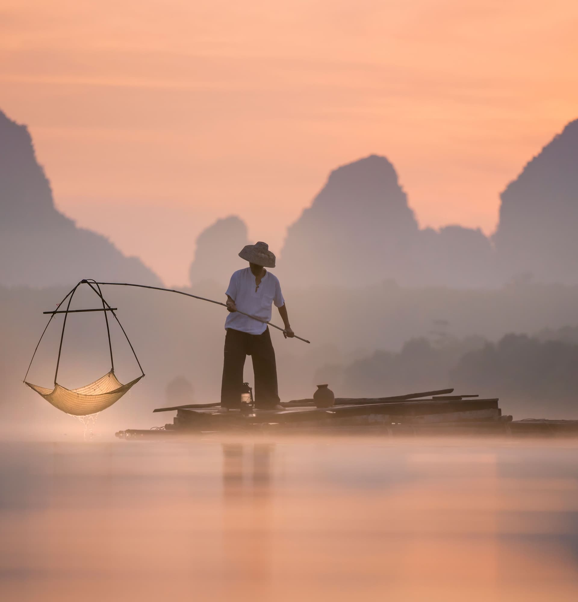 a Vietnamese fisherman on the water, holding a makeshift net above the water, as he stands on his wooden raft. The skyline is pink, with a beautiful mountainous backdrop.