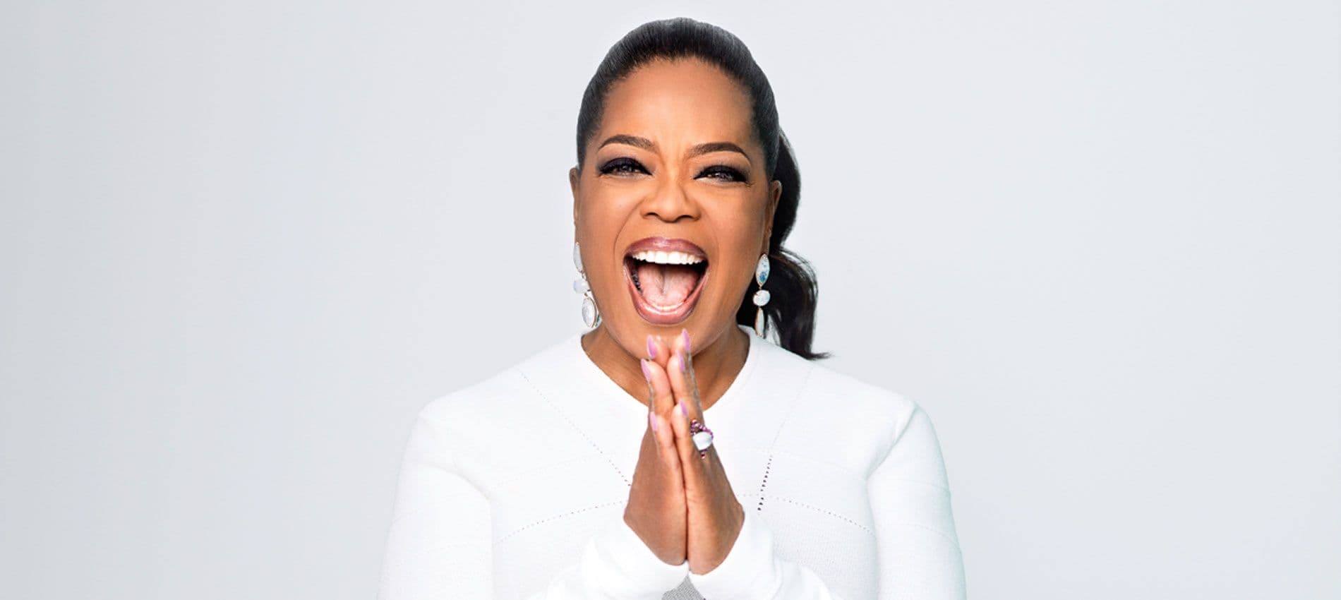Oprah wearing a white dress laughing with her hands pressed together.